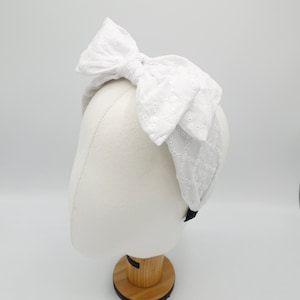 May include: White headband with a large, white, embroidered bow. The headband is made of a soft, stretchy fabric.