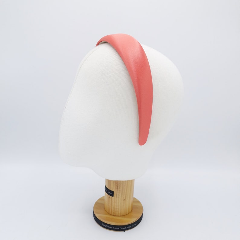 May include: A coral-colored headband with a smooth, matte finish. The headband is displayed on a white mannequin head, showcasing its shape and design. The headband is a simple, yet elegant hair accessory.