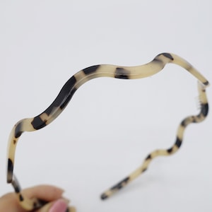 May include: A wavy headband with a tortoiseshell pattern. The headband features a light beige base with dark brown and black spots. The design is a continuous wave, adding a stylish touch to any hairstyle.