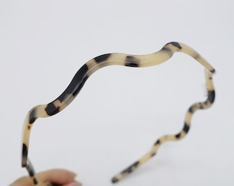Puede incluir: Una diadema ondulada con estampado de carey. El accesorio para el cabello presenta una base beige claro con manchas marrones oscuras y negras. El diseño ondulado añade un toque elegante a cualquier peinado.
