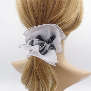 May include: A white and gray scrunchie with a layered, ruffled design. The scrunchie features a central, gathered section of gray velvet fabric, surrounded by layers of white and gray sheer fabric. The edges are trimmed with a thin gray border.