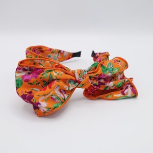 May include: A floral headband with a large orange bow. The bow is made of fabric with a floral print in shades of pink, purple, green, and white. The headband is black.