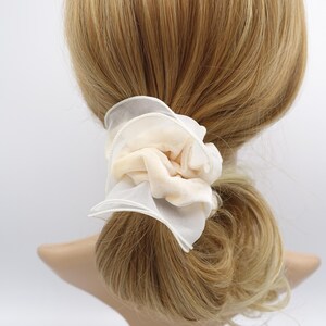 May include: A cream-colored scrunchie with a layered design. The scrunchie features a soft, velvety fabric in the center, surrounded by delicate, sheer white fabric. The hair accessory is tied around a ponytail.
