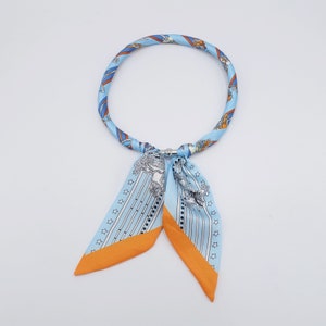 Könnte beinhalten: Ein hellblauer und orangefarbener Seidenschal mit Muster und einem Metallverschluss. Der Schal ist vorne zu einem Knoten gebunden und die Enden hängen herunter.