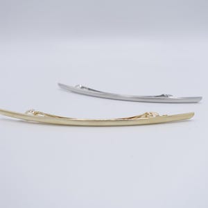 May include: Two curved metal hair barrettes, one in a polished gold tone and one in a silver tone. Each barrette features a sleek, elongated, slightly arched design with a functional spring-loaded clip mechanism visible on the underside.