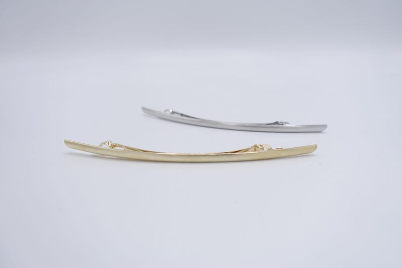 May include: Two curved metal hair barrettes, one in a polished gold tone and one in a silver tone. Each barrette features a sleek, elongated, slightly arched design with a functional spring-loaded clip mechanism visible on the underside.