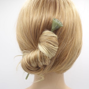 Peut inclure: Une épingle à cheveux en forme de fleur de lotus verte est insérée dans un chignon de cheveux blonds. L'épingle à cheveux est de couleur vert pâle et présente un motif floral détaillé. Les cheveux sont coiffés en chignon bas, avec des mèches lâches encadrant la nuque.
