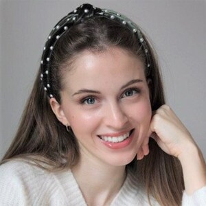 May include: A woman with long dark hair is wearing a black headband with pearl accents. The headband has a knot on top and is decorated with small white pearls.