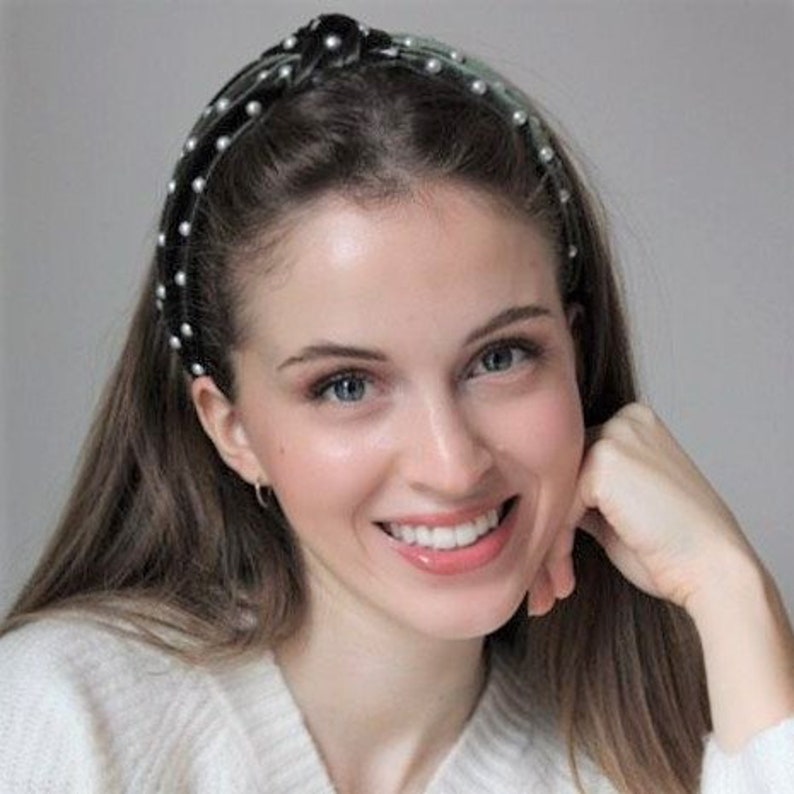 May include: A woman with long dark hair is wearing a black headband with pearl accents. The headband has a knot on top and is decorated with small white pearls.