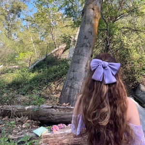 Peut inclure: Une femme avec de longs cheveux bruns portant une robe lavande et un grand nœud lavande dans les cheveux. Elle se tient dans une forêt avec un livre et des fleurs sur une bûche.