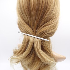 May include: A sleek, silver-toned metal hair barrette with a smooth, straight, and elongated design. The accessory is positioned horizontally across a section of light blonde hair, securing the strands in place with its minimalist, polished finish.