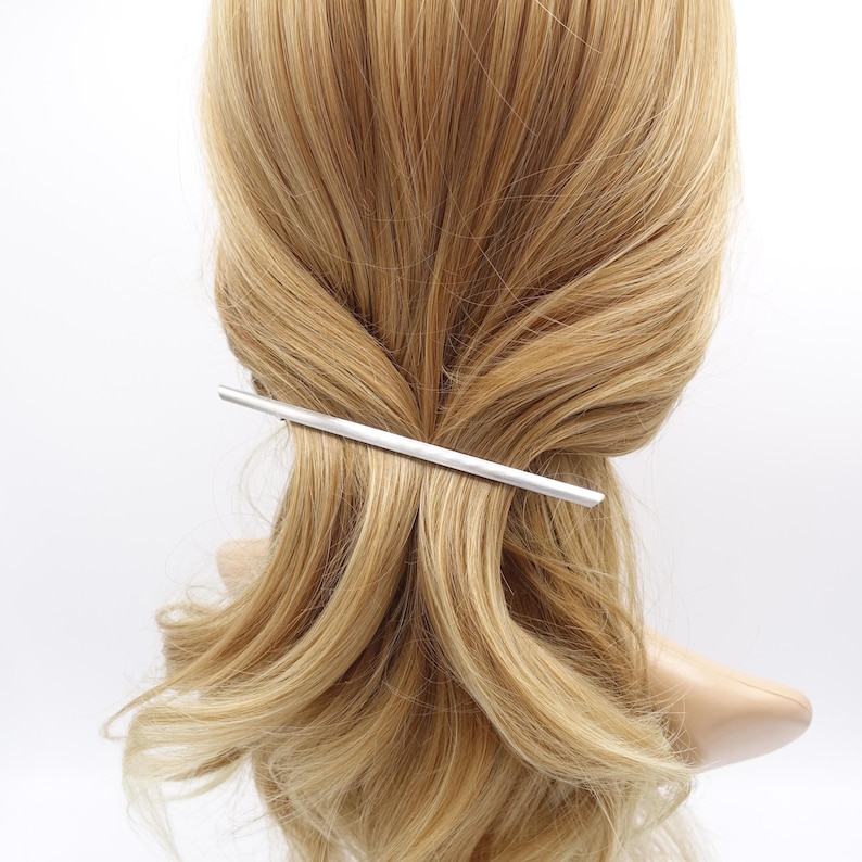 May include: A sleek, silver-toned metal hair barrette with a smooth, straight, and elongated design. The accessory is positioned horizontally across a section of light blonde hair, securing the strands in place with its minimalist, polished finish.
