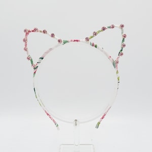 May include: A white headband with a floral print and pink rhinestones forming cat ears.