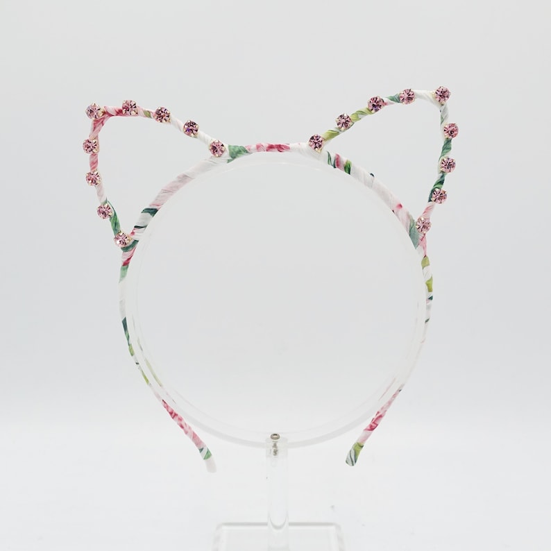 May include: A white headband with a floral print and pink rhinestones forming cat ears.