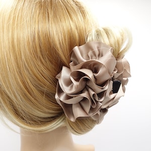 May include: A taupe satin hair clip shaped like a flower, with layered, ruffled petals. The hair accessory is attached to blonde hair, styled in a bun. The clip is a neutral color, suitable for various hairstyles.
