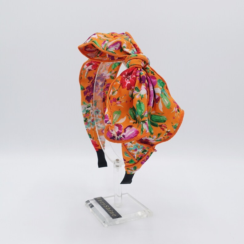 May include: Orange headband with a large floral bow. The bow is made of a fabric with a floral print in shades of pink, purple, green, and white. The headband is on a clear display stand.