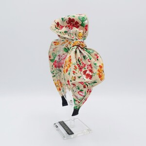 May include: A floral headband with a large bow. The headband is made of a beige fabric with a floral print in shades of red, yellow, and green. The bow is tied in a large, loose knot.