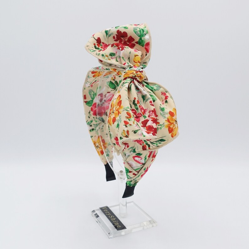 May include: A floral headband with a large bow. The headband is made of a beige fabric with a floral print in shades of red, yellow, and green. The bow is tied in a large, loose knot.