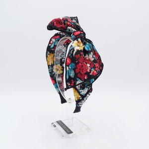 May include: A black headband with a large floral bow. The bow is made of a black fabric with a colorful floral print. The headband is on a clear display stand.