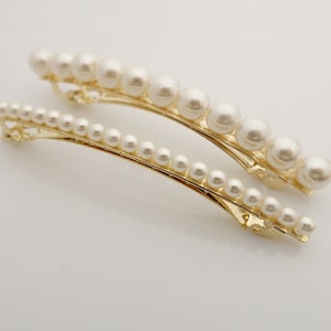 May include: Two gold hair clips with a row of faux pearls. The clips are curved and have a simple design.