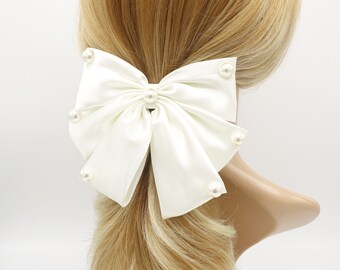 May include: A white satin bow with pearl embellishments. The bow is tied with a knot in the center and has a loop on each side. The bow is attached to a hair clip.