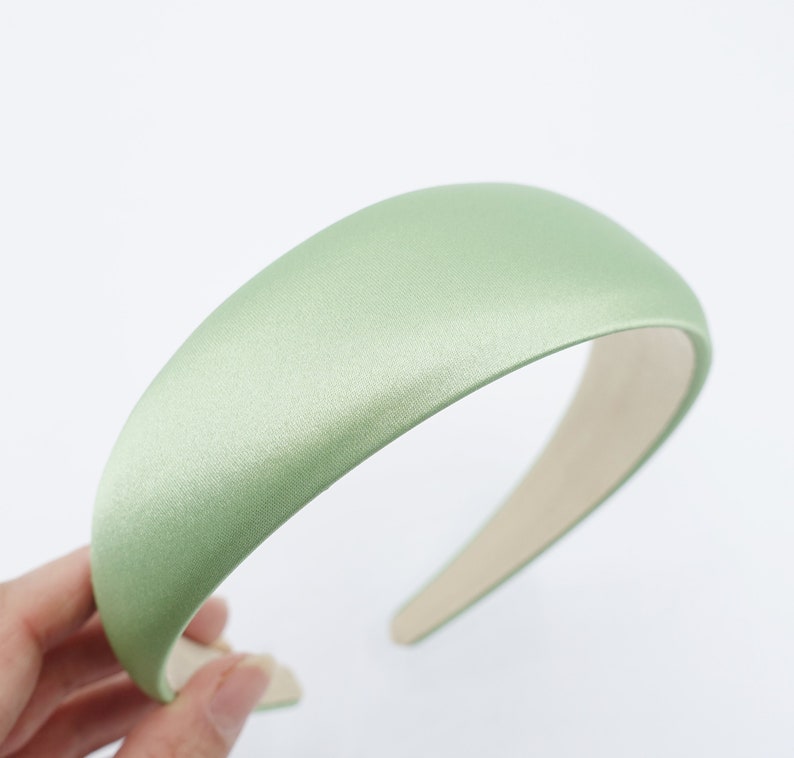 May include: A light green satin headband with a padded, rounded top. The headband is a solid color and has a smooth texture. The interior is a light beige color. The headband is a hair accessory.