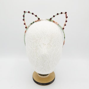 May include: A floral headband with cat ears adorned with burgundy rhinestones. The headband is displayed on a white lace-covered mannequin head.