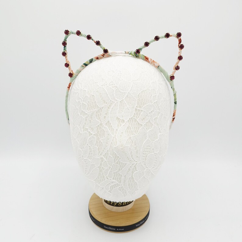 May include: A floral headband with cat ears adorned with burgundy rhinestones. The headband is displayed on a white lace-covered mannequin head.
