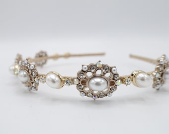 Puede incluir: Una diadema dorada con diseños florales y ovalados ornamentados. La diadema presenta perlas de imitación y pedrería transparente e iridiscente. Los elementos decorativos están colocados sobre una fina banda beige, creando un accesorio delicado y elegante.