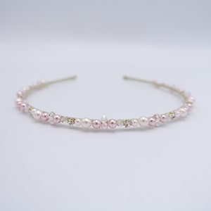 Puede incluir: Una diadema fina de metal en tono dorado adornada con una delicada disposición de pequeñas perlas redondas de color rosa claro y blanco, intercaladas con diminutos cristales transparentes y brillantes que captan la luz a lo largo de la banda.