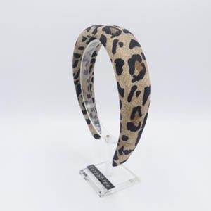 Puede incluir: Una diadema acolchada con estampado de leopardo. La diadema presenta un fondo beige con manchas negras. El accesorio se exhibe sobre un soporte transparente sobre un fondo blanco. El texto "Very Shine" es visible en el soporte.