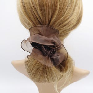 May include: A brown scrunchie with a ruffled, sheer outer layer and a velvet-like center. The hair accessory is wrapped around a ponytail of blonde hair, creating a soft, textured look. The scrunchie adds a touch of elegance to any hairstyle.