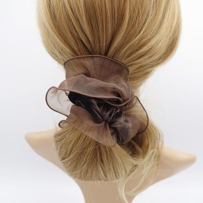 May include: A brown scrunchie with a ruffled, sheer outer layer and a velvet-like center. The hair accessory is wrapped around a ponytail of blonde hair, creating a soft, textured look. The scrunchie adds a touch of elegance to any hairstyle.