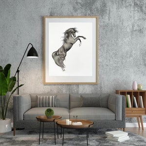 May include: A framed black and white drawing of a rearing horse. The artwork is displayed in a room with a gray sofa, wooden side tables, and a floor lamp. The horse is depicted with detailed shading, showcasing its muscular form.