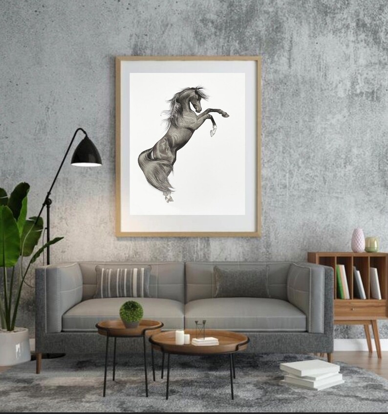 May include: A framed black and white drawing of a rearing horse. The artwork is displayed in a room with a gray sofa, wooden side tables, and a floor lamp. The horse is depicted with detailed shading, showcasing its muscular form.