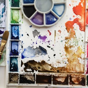 May include: A watercolor artist's palette with various colors, including blues, greens, reds, and browns. The palette is white with multiple wells and a circular mixing area. Two paint brushes are visible, one with a red handle.