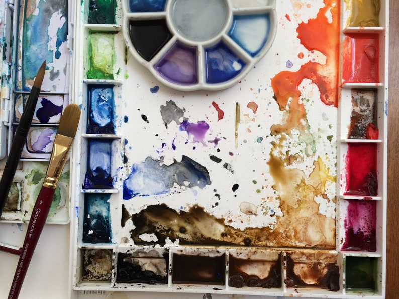 May include: A watercolor artist's palette with various colors, including blues, greens, reds, and browns. The palette is white with multiple wells and a circular mixing area. Two paint brushes are visible, one with a red handle.