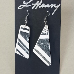 May include: A pair of silver earrings with a black and white striped design. The earrings are hanging from a black card with the text "L Henry" written in silver.