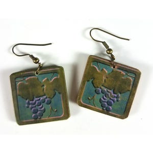 May include: A pair of square-shaped earrings with a grapevine design. The earrings are made of a light brown material with a green and blue glaze. The grapevine design is in a darker brown color. The earrings have a bronze-colored hook.