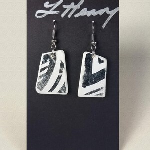 May include: A pair of white rectangular earrings with black and white geometric designs. The earrings are hanging from silver hooks.
