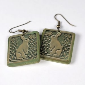 May include: A pair of green ceramic earrings with a raised image of a rabbit in a field of grass. The earrings have bronze hooks.