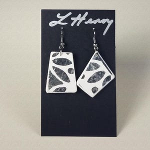 May include: A pair of white earrings with a black and white geometric pattern. The earrings are hanging from a black card with the text "L'Henry" written in silver.