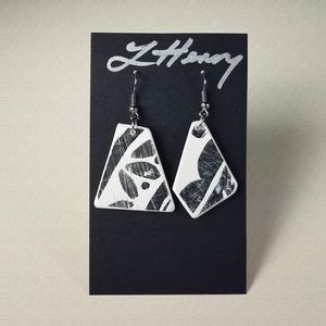 May include: A pair of white ceramic earrings with black geometric designs. The earrings are hanging from a black card with the word "L'Honey" written in silver lettering.