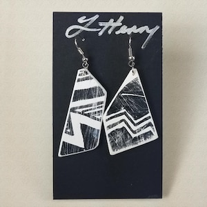 May include: A pair of white geometric earrings with black geometric designs. The earrings are hanging from silver hooks on a black background.