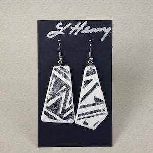 May include: A pair of white ceramic earrings with a black geometric pattern. The earrings are hanging from silver hooks on a black card with the signature "L Henry" written in silver.