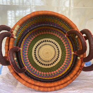 May include: A round, hand-woven basket featuring a vibrant, multi-coloured geometric pattern in orange, green, purple, and cream. The basket includes two sturdy, dark brown leather-wrapped handles attached to the sides for easy carrying.