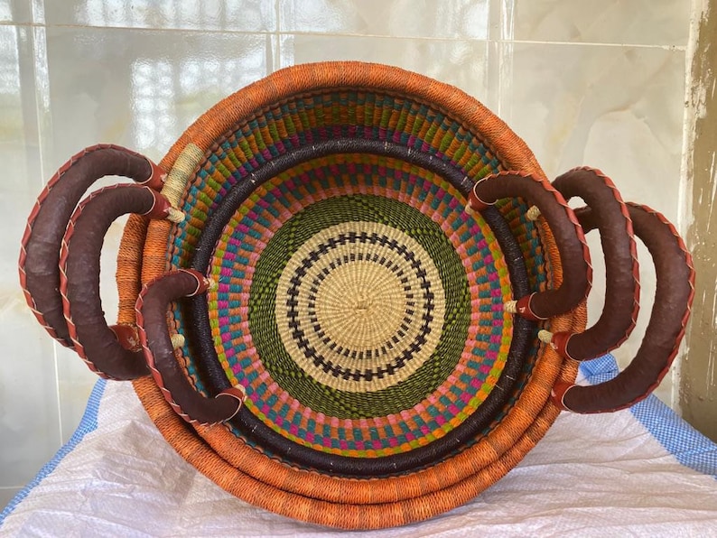 May include: A round, hand-woven basket featuring a vibrant, multi-coloured geometric pattern in orange, green, purple, and cream. The basket includes two sturdy, dark brown leather-wrapped handles attached to the sides for easy carrying.