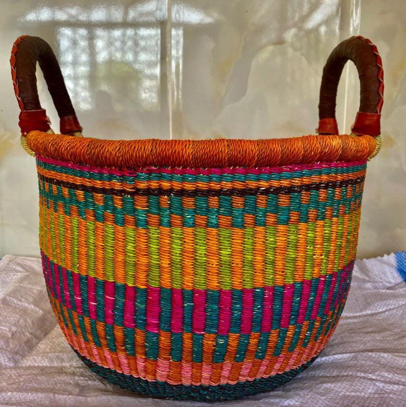 May include: A round woven basket featuring horizontal bands of vibrant orange, teal, lime green, and hot pink. The basket has two sturdy, dark brown leather-wrapped handles attached to the top rim, which is finished with a thick orange coil.