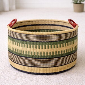 Op de afbeelding: Een ronde geweven opbergmand met een beige, groen en marineblauw gestreept patroon. De mand heeft rode leren handvatten aan elke kant. De mand is gemaakt van natuurlijke materialen en is geschikt voor woondecoratie.