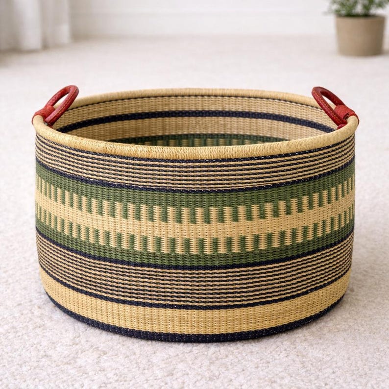 Op de afbeelding: Een ronde geweven opbergmand met een beige, groen en marineblauw gestreept patroon. De mand heeft rode leren handvatten aan elke kant. De mand is gemaakt van natuurlijke materialen en is geschikt voor woondecoratie.